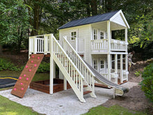 Load image into Gallery viewer, Two-story white wooden playhouse with double balconies, a slide, and climbing wall, nestled in a forest garden setting by WholeWoodPlayhouses