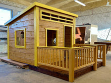 Load image into Gallery viewer, Wooden playhouse with yellow trim in a workshop setting, terrace and railings