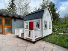 Load image into Gallery viewer, White farmhouse-style wooden playhouse with a red door, black metal roof, and a small front porch, assembled in the Colorado mountains. The playhouse is available for sale at Whole Wood Playhouses
