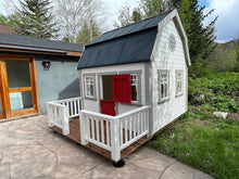Load image into Gallery viewer, White farmhouse-style wooden playhouse with a red door, black metal roof, and a small front porch, assembled in the Colorado mountains. The playhouse is available for sale at Whole Wood Playhouses