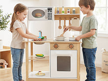 Load image into Gallery viewer, Two children playing with a wooden toy kitchen set in a bright room. The play kitchen features a white oven, stovetop, shelves with toy food and bottles, and hanging utensils.