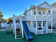 Load image into Gallery viewer, Second close-up of the kids playhouse, left and front. White two-storey kids playhouse with black metal roof, terrace on the ground floor and balcony with roof on the second floor. Flower boxes on balcony railings. Flower boxes on the first floor railings have red flowers. Balcony to the side, blue plastic slide and wooden climbing wall. Under the balcony is a sandpit. Artificial grass around the playhouse. By WholeWoodPlayhouses
