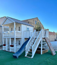 Load image into Gallery viewer, Right and front view of the kids playhouse. White two-storey kids playhouse with black metal roof, terrace on the ground floor and balcony with roof on the second floor. Flower boxes on balcony railings. Flower boxes on the first floor railings have red flowers. Balcony, slide and climbing wall on the side. Under the balcony is a sandpit. Artificial grass around the playhouse. By WholeWoodPlayhouses