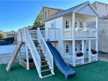 Load image into Gallery viewer, Left and foreground view of the kids playhouse. White two-storey kids playhouse, with terrace on the ground floor and balcony with roof on the second floor. Balcony, slide and climbing wall on the side. Under the balcony is a sandpit. By WholeWoodPlayhouses
