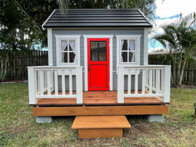 Load image into Gallery viewer, Children's playhouse Kids Cave façade image, red wooden door with safety glass window, grey walls, white safety glass opening windows, black steel roof, white decking railing and natural wood colored terrace by WholeWoodPlayhouses.