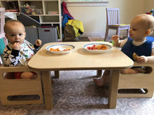 Load image into Gallery viewer, Two children sitting at a small wooden table with plates of food in a home setting.