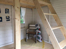 Load image into Gallery viewer, Interior view of a white wooden playhouse with kids chair, pictures on the wall and ladder  to the loft by WholeWoodPlayhouses