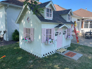 White Wooden Farmhouse Style Kids Outdoor Playhouse DIY Kit Little Farmhouse With pink door and window shutters, pink flower boxes with flowers in the Backyard by WholeWoodPlayhouses