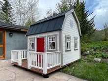Load image into Gallery viewer,  Outdoor Playhouse Farmhouse with black metal roof, a round top window and wooden terrace white wooden railing in the backyard by WholeWoodPlayhouses
