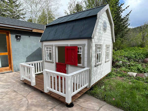 Kids Playhouse Farmhouse with  red dutch door, 4 opening windows and black metal roof in the backyard by WholeWoodPlayhouses