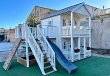 Load image into Gallery viewer, Close-up of the kids playhouse, left and front. White two-storey kids playhouse with black metal roof, terrace on the ground floor and balcony with roof on the second floor. Flower boxes on balcony railings. Flower boxes on the first floor railings have red flowers. Balcony to the side, blue plastic slide and climbing wall. Under the balcony is a sandpit. Artificial grass around the playhouse. By WholeWoodPlayhouses

