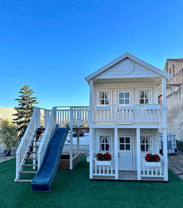 Front view, close-up of the kids playhouse, left and front. White two-storey kids playhouse with black metal roof, terrace on the ground floor and balcony with roof on the second floor. Flower boxes on balcony railings. Flower boxes on the first floor railings have red flowers. Balcony to the side, blue plastic slide and wooden climbing wall. Windows have Under the balcony is a sandpit. All opening windows have white curtains. Artificial grass around the playhouse. By WholeWoodPlayhouses