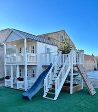Load image into Gallery viewer, Right and front view of the kids playhouse. White two-storey kids playhouse with black metal roof, terrace on the ground floor and balcony with roof on the second floor. Flower boxes on balcony railings. Flower boxes on the first floor railings have red flowers. Balcony, slide and climbing wall on the side. Under the balcony is a sandpit. Artificial grass around the playhouse. By WholeWoodPlayhouses

