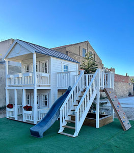 Right and front view of the kids playhouse. White two-storey kids playhouse with black metal roof, terrace on the ground floor and balcony with roof on the second floor. Flower boxes on balcony railings. Flower boxes on the first floor railings have red flowers. Balcony, slide and climbing wall on the side. Under the balcony is a sandpit. Artificial grass around the playhouse. By WholeWoodPlayhouses