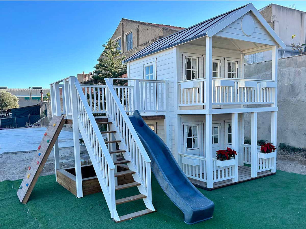 Left and foreground view of the kids playhouse. White two-storey kids playhouse, with terrace on the ground floor and balcony with roof on the second floor. Balcony, slide and climbing wall on the side. Under the balcony is a sandpit. By WholeWoodPlayhouses