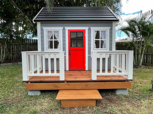 Children's playhouse Kids Cave façade image, red wooden door with safety glass window, grey walls, white safety glass opening windows, black steel roof, white decking railing and natural wood colored terrace by WholeWoodPlayhouses.