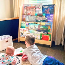 Load image into Gallery viewer, Two children reading books on the floor next to a wooden bookshelf filled with colorful books.
