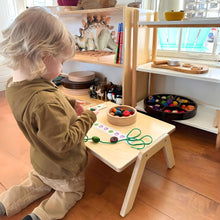 Load image into Gallery viewer, Child playing with educational toys at a wooden table in a room with shelves and windows.
