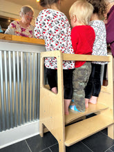 Load image into Gallery viewer, Children using a wooden step stool to reach a counter, with an adult nearby.
