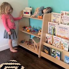 Load image into Gallery viewer, Child looking toys on a bookshelf against a blue-green wall
