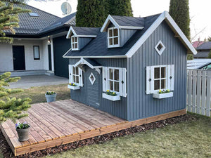 Outside Assembled Wooden Farmhouse Style Kids Outdoor Playhouse DIY Kit Little Farmhouse With Gray Walls, White Windows And Brown Terrace Next To Backyard White Fence by WholeWoodPlayhouses