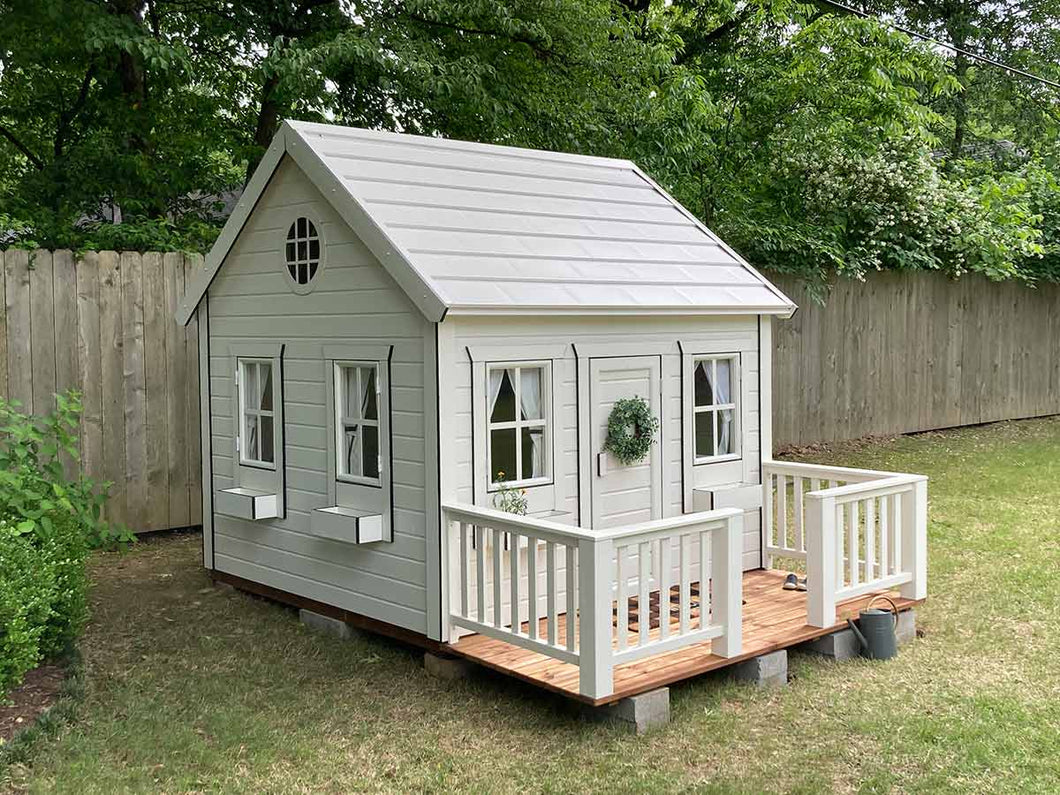 White Outdoor Kids Playhouse Snowy Owl With White Metal Roof, Brown Color Wooden Terrace and White Wooden Railing on Green Lawn Next To Gray Fence by WholeWoodPlayhouses 