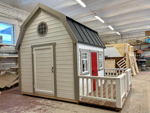 Kids OutDoor Playhouse Farmhouse Left Side View, With White Walls and Black Metal Roof, Terrace and White Railings, Beige Wall Trim, Dark Red Fishbone Style Wooden Dutch Door, One Full Size Adult Door, Two Front Side Opening and One Round Window in WholeWoodPlayhouses Shop