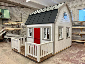 Kids OutDoor Playhouse Farmhouse Right Side View, With White Walls and Black Metal Roof, Terrace and White Railings, Beige Wall Trim, Dark Red Fishbone Style Wooden Dutch Door, Four Opening and One Round Window in WholeWoodPlayhouses Shop