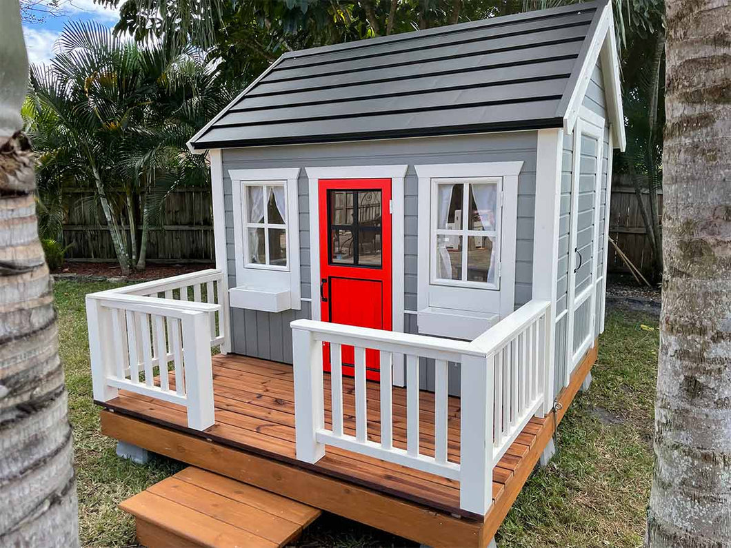 Wooden Playhouse Boy Cave With Black Metal Roof, a terrace with white railing and Red Half Glass Door in the backyard By WholeWoodPlayhouses